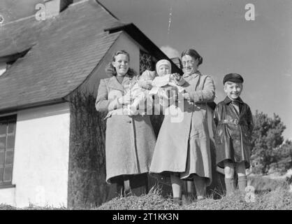 Evacuees To a Communal Hostel in Thurlestone, South Devon, England, 1941 Mrs Findlayson (left, with her 2-month-old daughter Sheila), and Mrs Phillips (with her sons, 9-month-old Roland and 6-year-old Roy) smile for the camera in the sunshine outside the 'Love Nest' in Thurlestone, Devon. Mrs Findlayson and Mrs Phillips have been evacuated to this communal hostel and are caretakers of this rest home, attached to the hostel, which is used by husbands visiting their wives at the Communal Billets whilst home on leave. Stock Photo