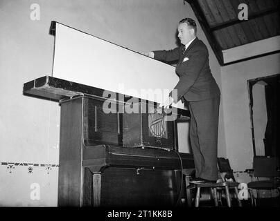 Film Show at Highest Village in the Highlands- Ministry of Information Film Screening, Tomintoul, Banffshire, Scotland, UK, 1943 Ministry of Information Film Operator John Macdougall sets up a screen and loudspeaker in the Memorial Hall in Tomintoul in preparation for the screening of a Ministry of Information film. The loud speaker is resting on the closed lid of an upright piano, with the screen, which Mr Macdougall is unfurling from its holder, resting on the top of the piano. Stock Photo