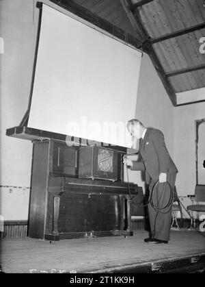 Film Show at Highest Village in the Highlands- Ministry of Information Film Screening, Tomintoul, Banffshire, Scotland, UK, 1943 Ministry of Information Film Operator John Macdougall sets up a screen and loudspeaker in the Memorial Hall in Tomintoul in preparation for the screening of a Ministry of Information film. The loud speaker is resting on the closed lid of an upright piano, with the screen resting on the top of the piano. Mr Macdougall is connecting the 'soundbox'. The original caption states that 'power is provided by the generator in [his] van outside'. Stock Photo