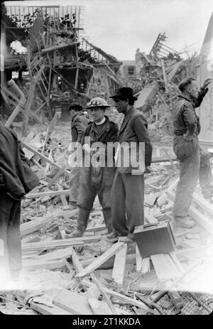 Flying Bomb- V1 Bomb Damage in London, England, UK, 1944 A vicar who has been helping with the rescue effort following a devastating V1 attack on the Highland Road and Lunham Road area of Upper Norwood, pauses in his work to smoke a cigarette and chat to one of his parishioners. The vicar is wearing overalls and a steel helmet. He also appears to have a pair of motorcycle goggles resting on the brim of his hat. They, and the Civil Defence workers around them, are standing on piles of rubble and timbers. Behind them, the devastated remains of several houses can be seen. Stock Photo