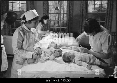Foster Parents Plan London Nursery- Caring For Displaced Children, London, England, 1941 Changing time in the 'big babies' room' at the 'Foster Parents Plan for War Children' Nursery in Hampstead. In the background, a German refugee woman who works at the home is changing the nappy of her own baby. In the foreground, one nurse changes a nappy, whilst a second, (on the right) cuts another baby's finger nails. Stock Photo