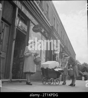 All-in War Worker- Everyday Life For Mrs M Hasler, Barnes, Surrey, 1942 Mrs Hasler, shopping baskets in hand, checks her ration book as she leaves a shop. Two other women can be seen in the photograph outside this small row of shops: one is a young mother with a pram, the other is carrying a basket and admiring the baby. Stock Photo