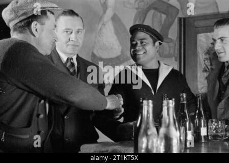 The Seven Seas Club- Life at the Merchant Navy Club, Edinburgh, Scotland, 1943 American seaman J C Winaski from Connecticut (left) shakes hands with Teaka Makario Bou, a Fighting French sailor from French Tahiti at the bar of the Seven Seas Club in Edinburgh. Andrew Jacobsen, from Norway, stands between them. Stock Photo