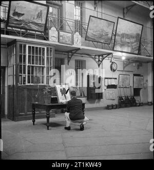 Wakefield Training Prison and Camp- Everyday Life in a British Prison, Wakefield, Yorkshire, England, 1944 An inmate crouches at a table to complete his painting in the central hall of Wakefield Training Prison. The hall forms the basis of the prison and has been decorated with paintings and drawings created by the prisoners in their spare time. Stock Photo