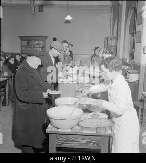 Woolmore Street Restaurant- Eating Out in Wartime London, 1942 The Woolmore Street British Restaurant works on a self-service basis, with diners taking their plates to the counter to be served. Here we see 77 year old T Hambly receiving his lunch from one of the 'dinner ladies', whilst others queue up behind him, receiving their choice of dish from the servers along the line. Mr Hambly was a merchant seaman during the First World War. Stock Photo