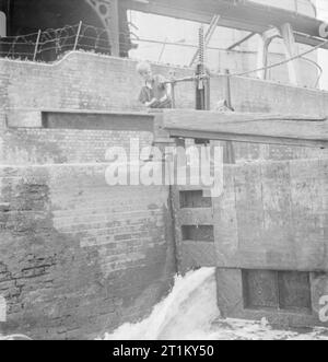 British Canals in Wartime- Transport in Britain, 1944 A young boy, son of a canal boatman, turns the handle of a lock, probably somewhere along Regent's Canal. He is winding up the paddles to let the water from the top level fill the lock and raise the boats. Stock Photo