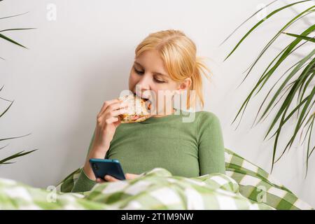 Languishing in bed, a girl faces mental decay, glued to her phone, and nibbling on toast throughout the day. Bed rotting Stock Photo