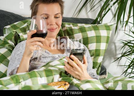 Engrossed in her phone, a middle-aged woman with short hair spends the entire day in bed, sipping wine. Inertia, life slowdown, lack of activity Stock Photo