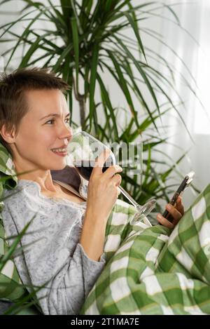 A woman in her middle years, sporting a short haircut, spends her entire day in bed, glued to her phone, sipping wine. Sloth, life deceleration, doing Stock Photo