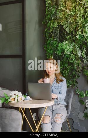 Woman working on laptop in cafe. Middle-aged freelancer with cup of coffee typing and reading. Loft working place with green plants Stock Photo