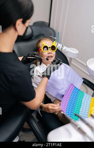Vertical view from shoulder of pediatric dentist examining and treats teeth of little child girl in funny eyeglasses lying on dental chair in Stock Photo
