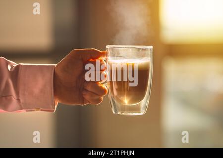 A cozy photo of a hand holding a steaming cup of coffee. The photo invokes warm feelings associated with sipping hot drinks before working day. Stock Photo