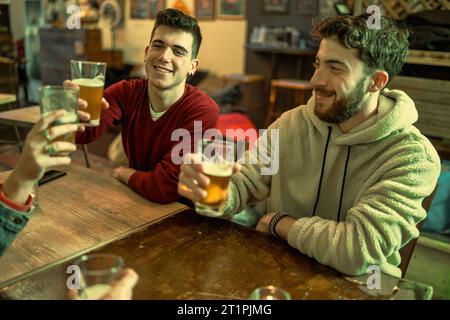 Two young men, clad in cozy attire, share a light-hearted moment as they raise their glasses in a toast at a rustic tavern. Stock Photo