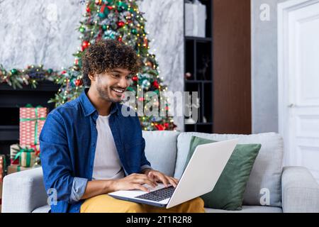 Young smiling man working remotely from home for Christmas sitting on couch in living room near decorated Christmas tree, Hispanic man using laptop, typing on keyboard. Stock Photo