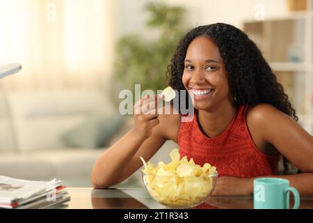 Happy black woman eating potato chips and posing looking at you at home Stock Photo