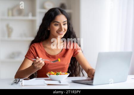 Smiling Woman Eating Lunch And Using Laptop At Home Office Stock Photo