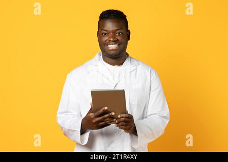 Cheerful black doctor man with tablet computer on yellow background Stock Photo