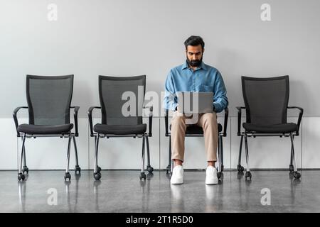 Indian man sitting on chair, waiting for interview and using laptop computer, full length, free copy space Stock Photo