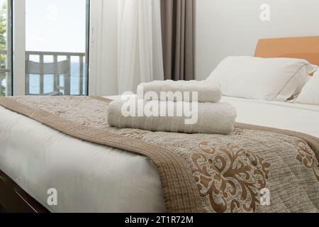 Double bedroom with double bed and sea view. White linen on bed, 2 pillows. Edge of bed is covered with brown bedspread, a stack of 2 white towels on Stock Photo
