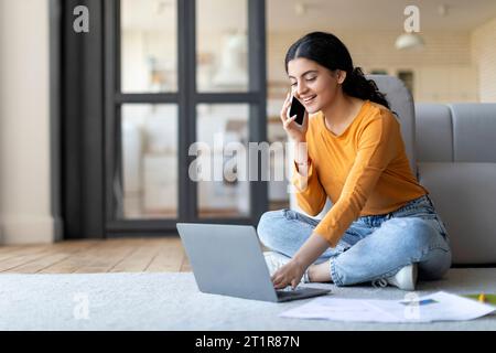 Young Indian Female Freelancer Using Laptop And Talking On Cellphone At Home Stock Photo