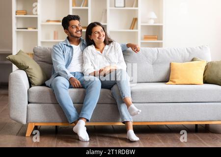 Happy dreamy indian couple looking aside at free space while sitting on sofa Stock Photo