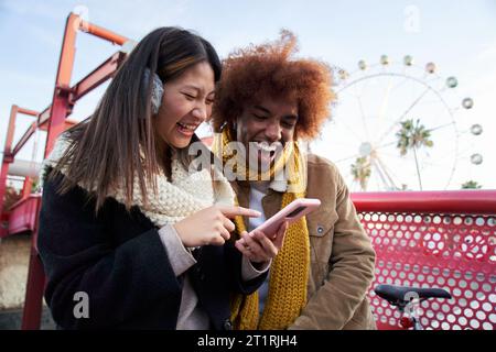 Multi-ethnic young couple friends in winter clothes looking laughing mobile outdoor. Amusement park. Stock Photo
