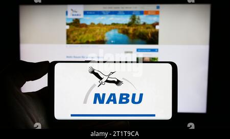 Person holding cellphone with logo of association Naturschutzbund Deutschland e.V. (NABU) in front of webpage. Focus on phone display. Stock Photo