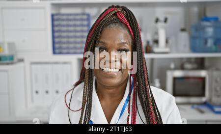 Confident african american woman scientist with braids, smiling beautifully in busy analytics chemistry lab, engrossed in medical research using micro Stock Photo