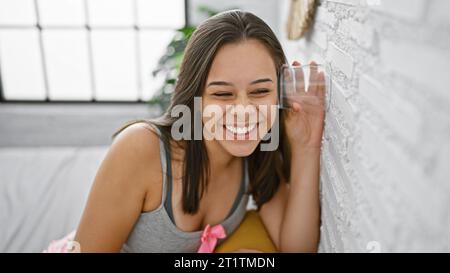 Intriguingly nosy young hispanic woman with attractive hairstyle, eavesdropping through bedroom wall with a glass, curious about private neighbour gos Stock Photo