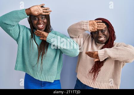 Two african woman standing over blue background smiling cheerful playing peek a boo with hands showing face. surprised and exited Stock Photo