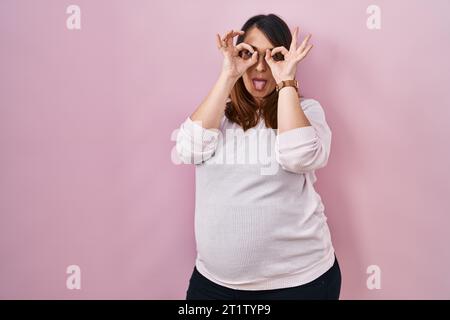 Pregnant woman standing over pink background doing ok gesture like binoculars sticking tongue out, eyes looking through fingers. crazy expression. Stock Photo