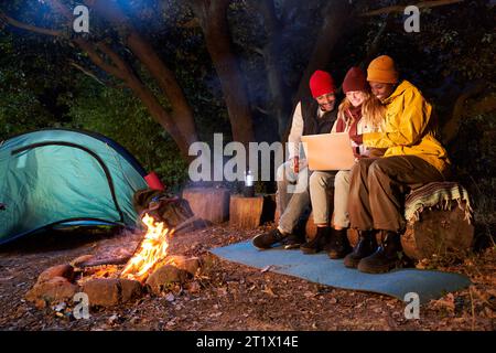 Three friends using laptop sitting together in front campfire one winter night. Happy young people. Stock Photo