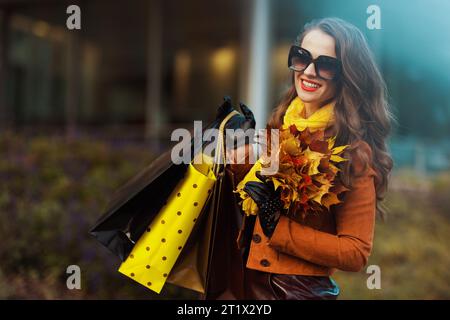 Hello autumn. smiling modern female in orange trench coat with shopping bags and autumn yellow leaves near shop in the city. Stock Photo