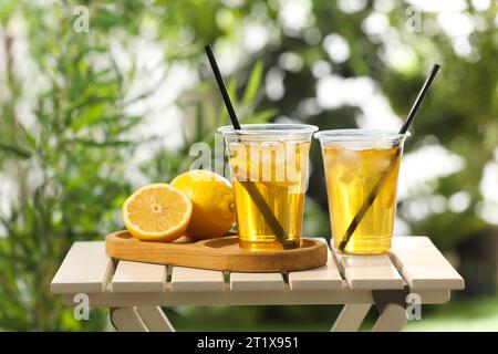 Plastic cups of tasty iced tea with lemon and fresh fruits on white wooden table against blurred green background Stock Photo