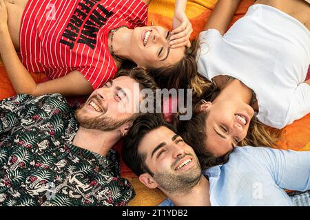 Four young adults, two men and two women, are seen from a top-down perspective, lying in a circle formation, smiling joyfully at the camera. Stock Photo