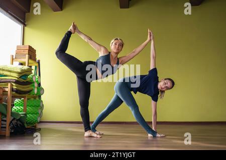 Two women yoga practitioners perform exercises Trikonasana, triangle pose and Natarajasana, king of dance pose, training in sportswear while standing Stock Photo