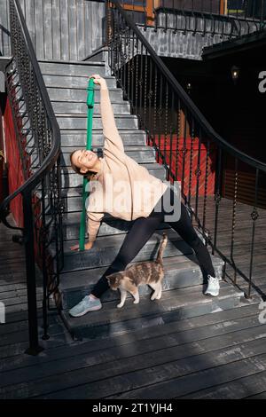 Satisfied young woman with her cat plays sports on the street, performs the Trikonasana exercise, triangle pose with a fitness belt, practices yoga in Stock Photo