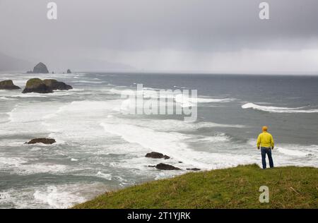 A young man, wearing a yellow jacket, stands on an overlook on the Oregon coast. Stock Photo
