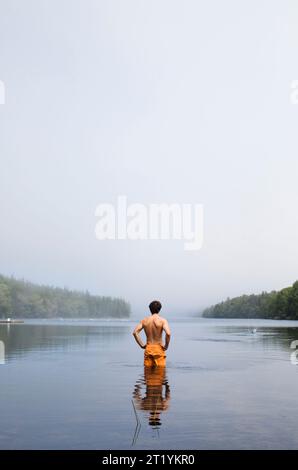 A young man stands in a bathing suit looking out over a small lake on a cloudy day. Stock Photo