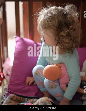 Toddler with pacifier holding baby doll in brown crib Stock Photo