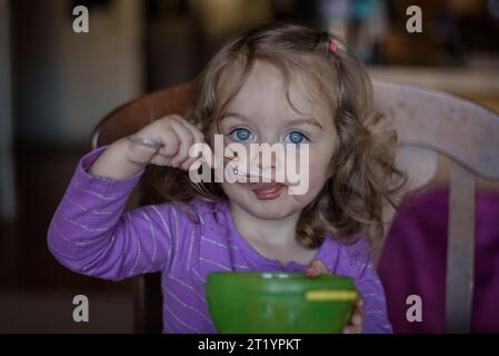 Beautiful little girl with big blue eyes and curls using spoon to eat Stock Photo