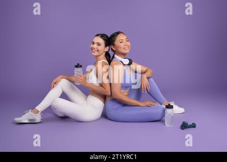 Diverse ladies friends resting with water bottles after workout, sitting on purple studio background Stock Photo