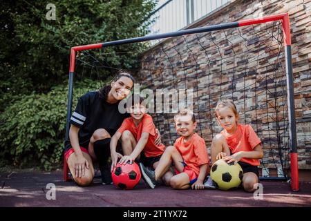 Mom playing football with her children, dressed in football jerseys. The family as one soccer team. Family sports activities outside in the backyard Stock Photo