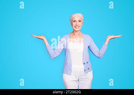 Beautiful nice cheerful smiling gray-haired old lady wearing casual, showing two options with palms. Isolated over pastel violet purple background Stock Photo
