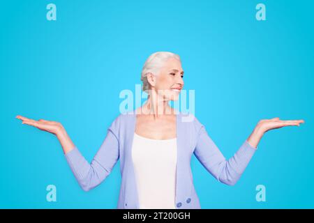 Beautiful nice cheerful smiling gray-haired old lady wearing casual, showing two options with palms, looking at one alternative. Isolated over pastel Stock Photo
