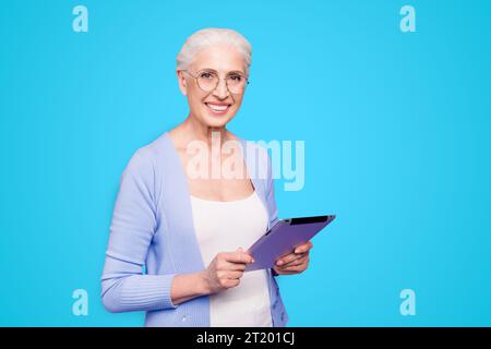 Grey haired old smiling business woman wearing glasses, reading browsing using holding tablet. Isolated over violet purple background Stock Photo