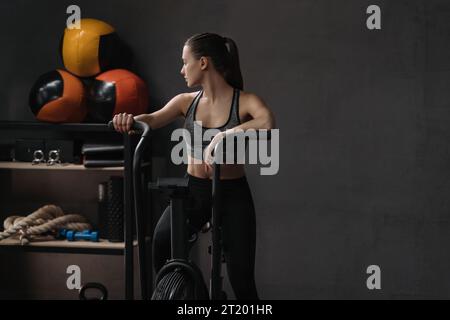 Portrait of female athlete on air bike at fitness gym. Sporty fit woman resting while doing cardio exercises on airbike, looking away. Copy space, gym background. Fitness and crossfit concept Stock Photo