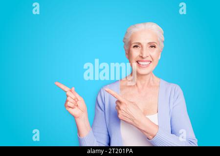Beautiful nice cheerful smiling gray-haired old lady wearing casual, showing pointing way with two fingers. Copy space. Isolated over pastel violet Stock Photo