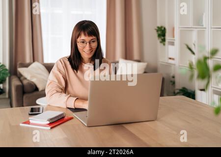 Pleasant happy young woman freelancer working on laptop at home. Attractive businesswoman in eyeglasses studying online, using laptop software, web surfing information or doing online shopping Stock Photo