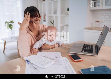 Unhappy frustrated single mother with crying children trying to work at home or solve family financial troubles, tired of balancing motherhood and work, doesn't have money to pay domestic bills Stock Photo